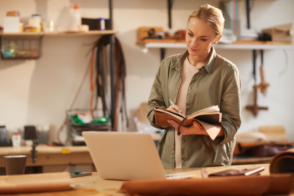 Kvinne som jobber i et verksted, skriver i en notatbok og bruker en laptop.